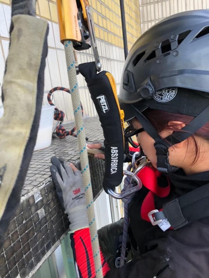 這張照片是一個正在高空作業的女性。她穿著安全帽、安全帶和手套,手裡握著一根繩索。她正在檢查建築物的外牆。