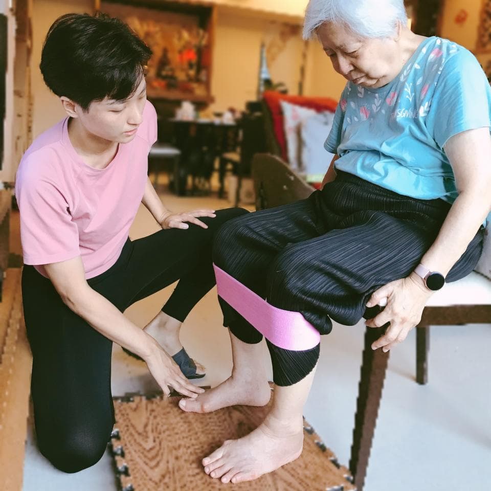 這張照片是一名年輕女性正在幫助一名老年女性做腿部運動,老年女性的腿上綁著一條粉色阻力帶。