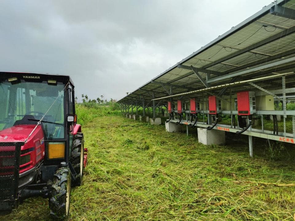 這張照片是一台紅色拖拉機停在太陽能板架旁邊,太陽能板架上面有許多太陽能板。