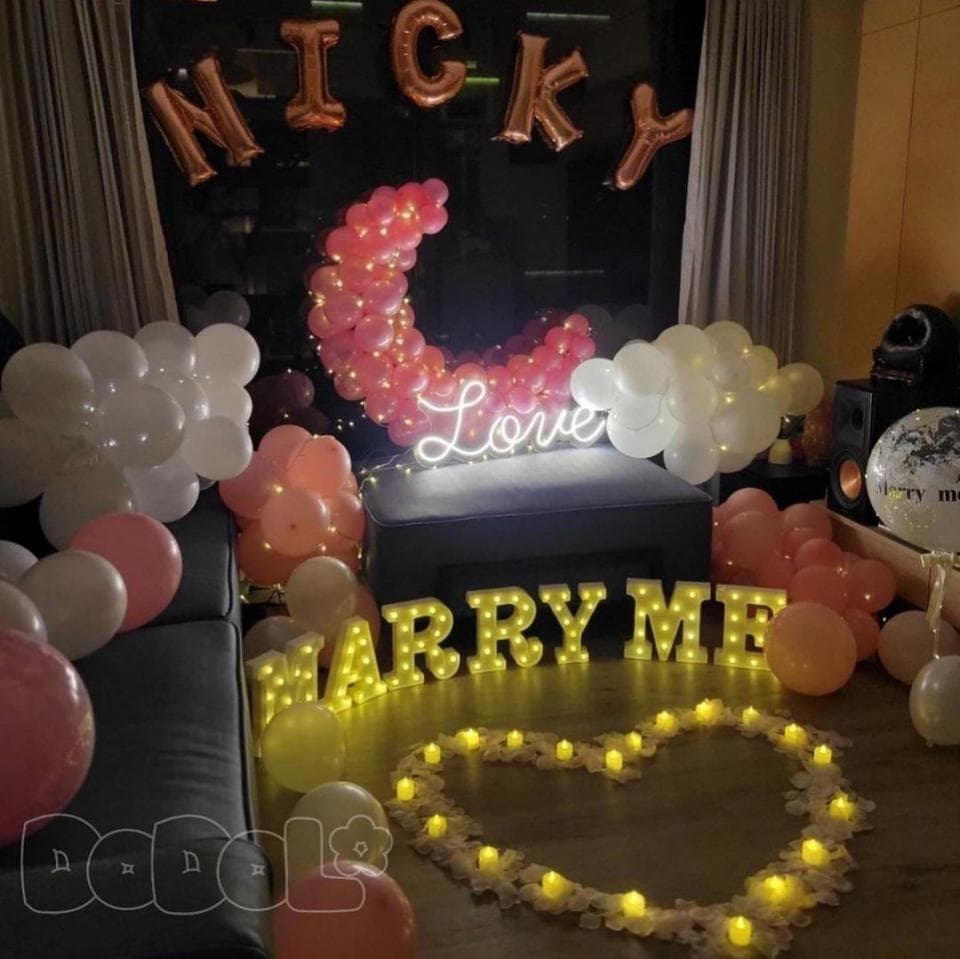 這張照片是一場浪漫的求婚佈置,最重要的是閃爍著金色光芒的「Marry Me」字樣。字樣旁圍繞著白色氣球和花瓣,營造出溫馨的氛圍,讓人感受到滿滿的愛意。