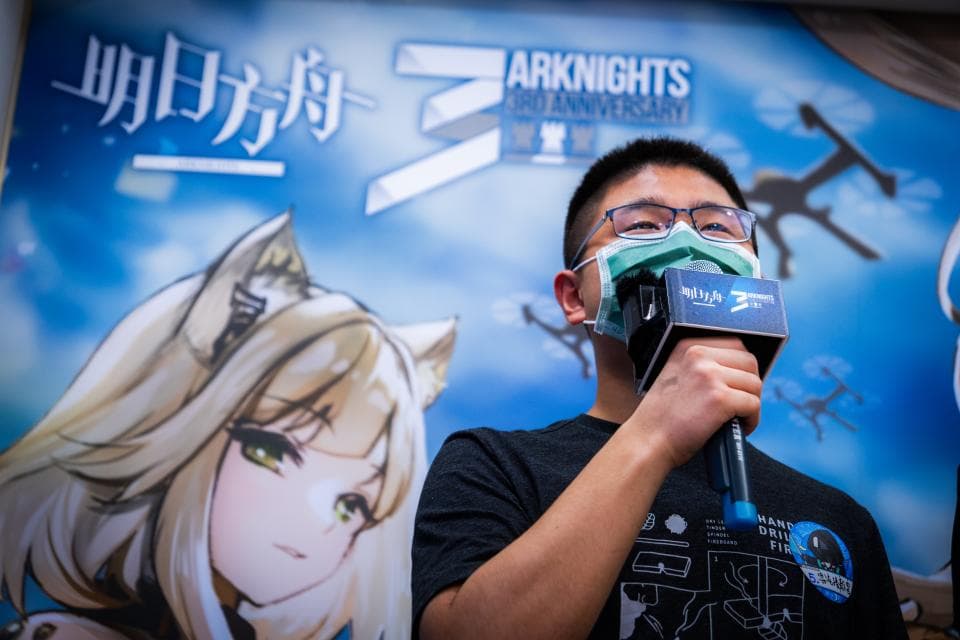 這張「明日方舟」的照片是一個男人正在拿著麥克風講話,他戴著口罩和眼鏡,後面是一幅明日方舟的宣傳畫。