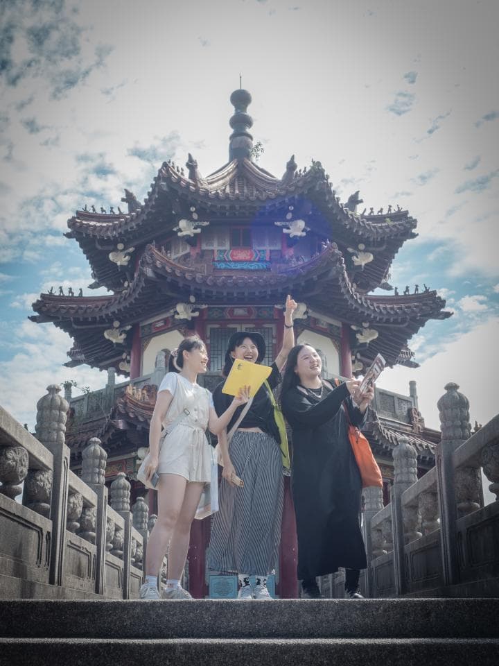 這張「三個女生在中國式建築前拍照」的照片是,三個女生站在一座中國式建築前,其中一個女生拿著黃色卡片,另一個女生則拿著手機自拍。她們的背後是一座有著紅色樑柱和屋簷的傳統建築,看起來像是寺廟或道觀。