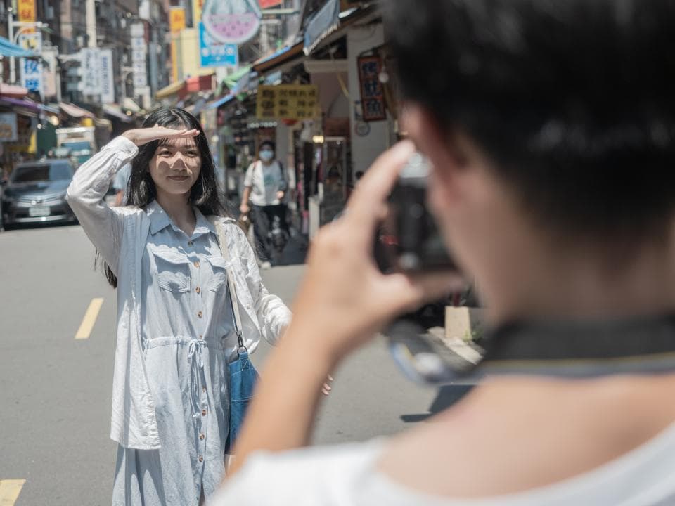 這張照片是一名女子在街道上被拍照,她穿著白色襯衫和藍色裙子,手遮陽,背景是街景。