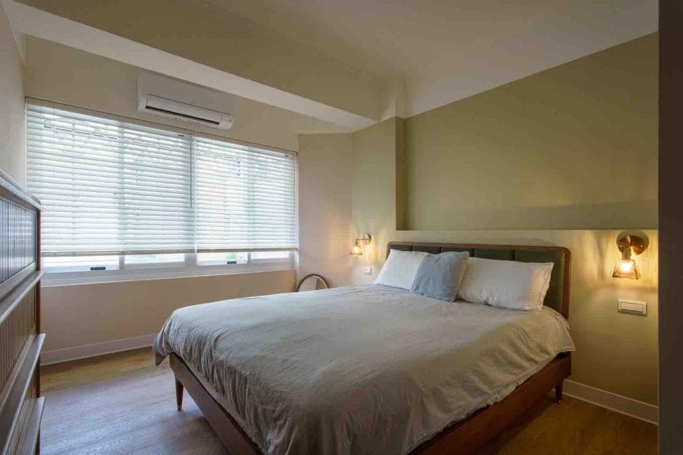 這張照片是一間臥室,房間裡有一張大床,床上鋪著白色床單,床頭有兩個床頭櫃,牆上掛著燈,窗戶上裝著百葉窗,房間整體看起來很舒適。