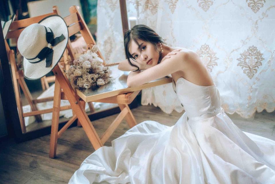 這張照片是一個穿著白色婚紗的新娘,她坐在一張木頭折疊椅上,看起來很優雅。她旁邊有一個裝飾花束的託盤,牆壁上有一個白色蕾絲的窗簾。