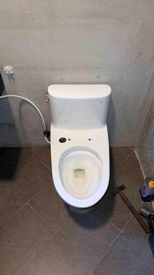 1) 照片顯示一個白色的馬桶,它安裝在一個灰色瓷磚地板上,地板上有工具和灰色的牆壁