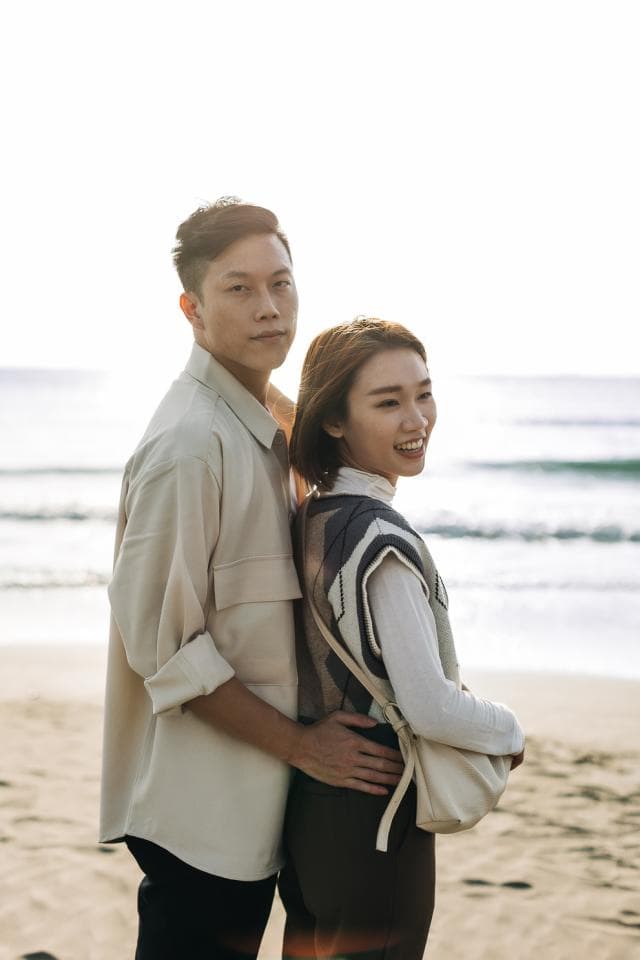 這張「情侶在海邊散步」的照片是,一個男人穿著米色的襯衫,摟著穿著白色毛衣的女人,他們都望向遠方,背景是海灘和藍天。