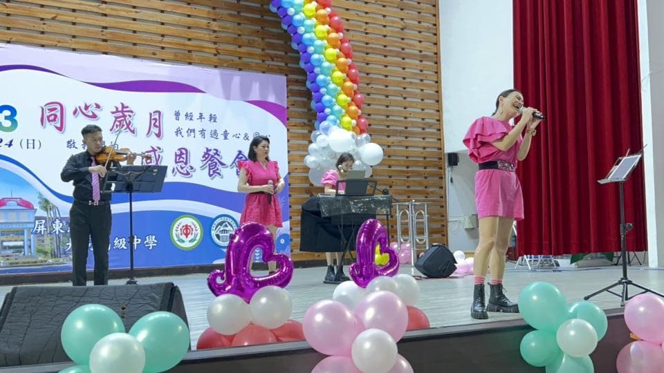 這張照片是舞臺表演,一個穿著粉色洋裝的女生正在唱歌,她身旁有其他人正在演奏樂器。