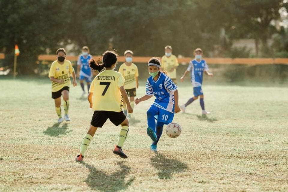 這張足球比賽的照片是 一位穿著藍色球衣的女孩正在控球,她的隊友穿著黃色球衣,他們正在草地上比賽。