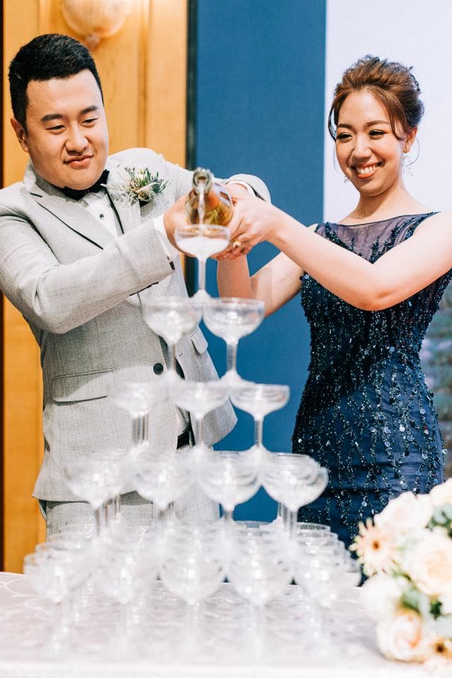 這張照片是一對新人舉杯慶祝的畫面,他們手中拿著香檳,背景是一座高聳的香檳塔,象徵著他們美好的未來。