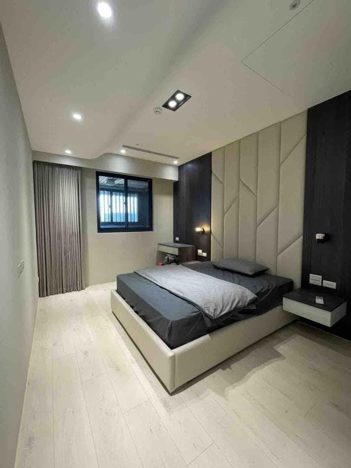 1) 一張照片顯示一個現代臥室,房間以白色和灰色為主色調,床鋪上有灰色床單和枕頭,床頭板是白色皮革,配有牆上的裝飾性縫線