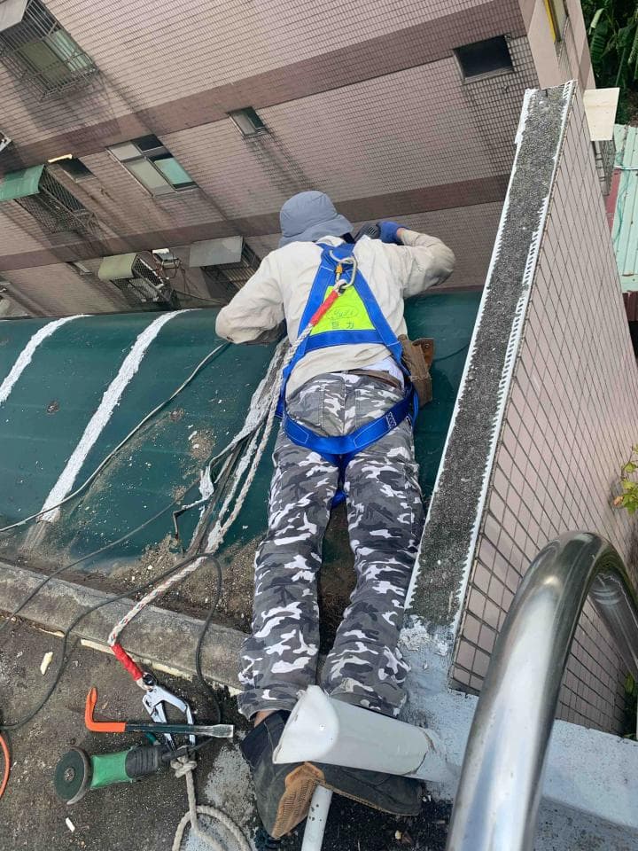 這張照片是一個工人正在屋頂上工作。他穿著安全帶和安全帽,並且正在使用工具來完成工作。他看起來很專注於他的工作,並且很小心地避免危險。