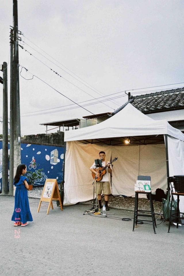 這張照片是一名男子在白色帳篷下彈吉他唱歌。他戴著帽子,穿著卡其色的褲子和白色襯衫,正在演奏一把棕色吉他。他面向前方,面帶微笑。