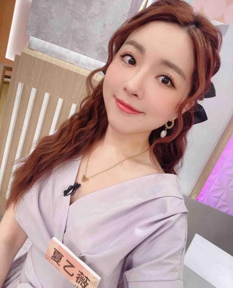 這張照片是一個穿著淺紫色上衣的女孩,她留著一頭波浪的棕色頭髮,並且戴著一條金項鍊和一對耳環。她的上衣有領口,而且有一個小標籤,上面寫著「真之福」。