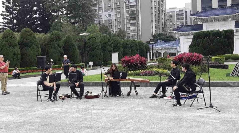 這張「戶外演奏」的照片是五個人在一個公園裡演奏音樂,其中一位女性彈奏古箏,兩位男性演奏吉他,另兩位則坐在椅子上欣賞音樂。