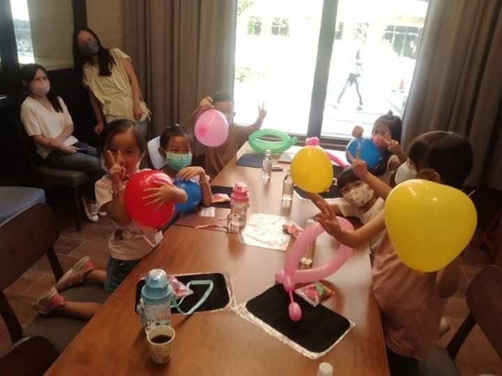1) 照片中顯示一群兒童在室內聚會,其中一些兒童拿著氣球玩耍,而其他兒童則坐在一張木桌上,桌上有氣球、飲料和其他物品