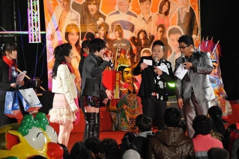 這張照片是一個慶祝活動,最重要的是舞臺上的兩位主持人,他們穿著正式服裝,一位手拿麥克風,另一位手拿卡片,看起來正在進行熱鬧的演講。