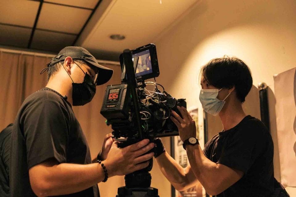 這張「電影拍攝」的照片是兩個戴著口罩的人,其中一個手持攝影機,另一個正在調整設備,拍攝過程中非常專注。