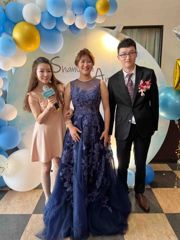 這張照片是一對新人結婚典禮的合照,照片中,新娘穿著一件深藍色的婚紗,裙擺蓬鬆,上面還有一些蕾絲花紋,新郎則穿著黑色西裝,打著領帶,兩個人都露出幸福的笑容,看起來非常恩愛。