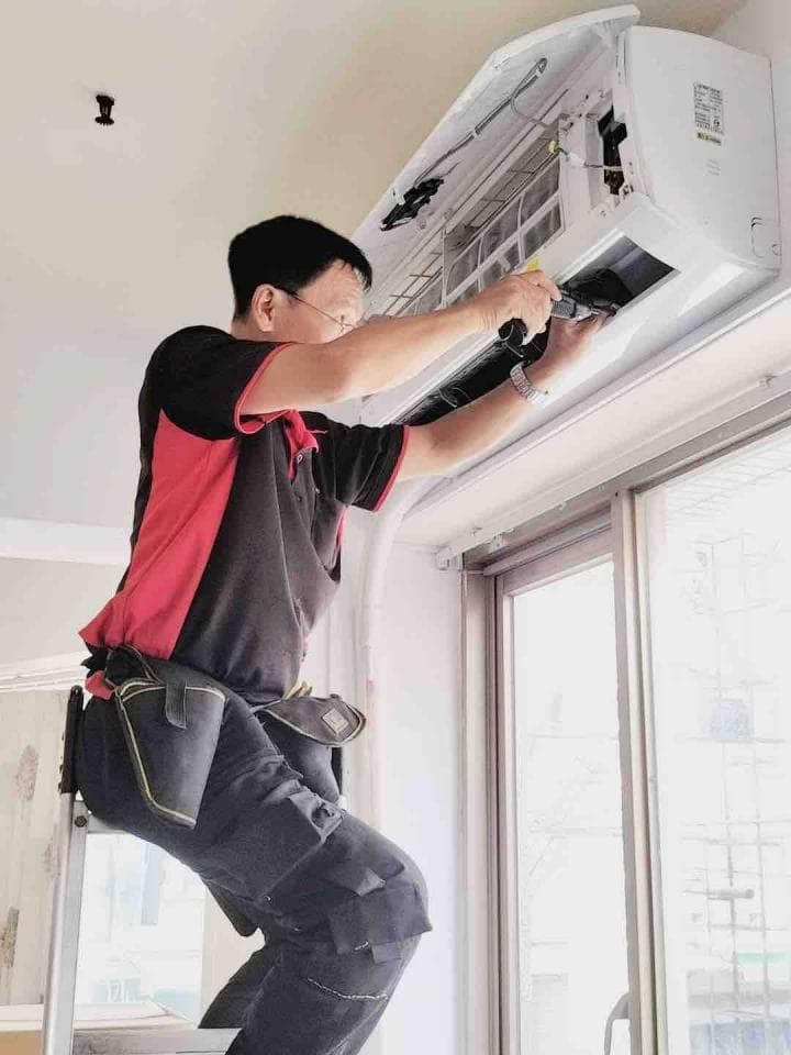 ## 照片描述
照片中,一名身穿紅黑條紋短袖上衣和黑色工作褲的男性正在修理安裝在牆上的冷氣機