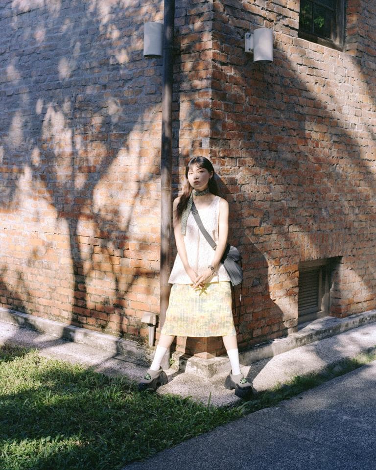 這張照片是一個穿著黃色短裙的女生站在紅磚牆邊。她穿著白色上衣,頭上綁著一條絲巾。她看起來很開朗,正在微笑著。