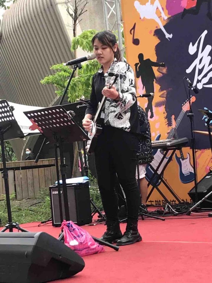 這張照片是一個女孩在舞臺上彈吉他的表演,女孩身穿黑色褲子和印花外套,正在認真地彈奏著吉他,她的專注神情和手中的吉他,是照片中最重要的部分。