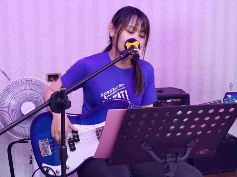 1) 照片中有一位年輕女子站在舞台上,穿著藍色 T 恤,拿著一把藍色的貝斯吉他,並用麥克風唱歌