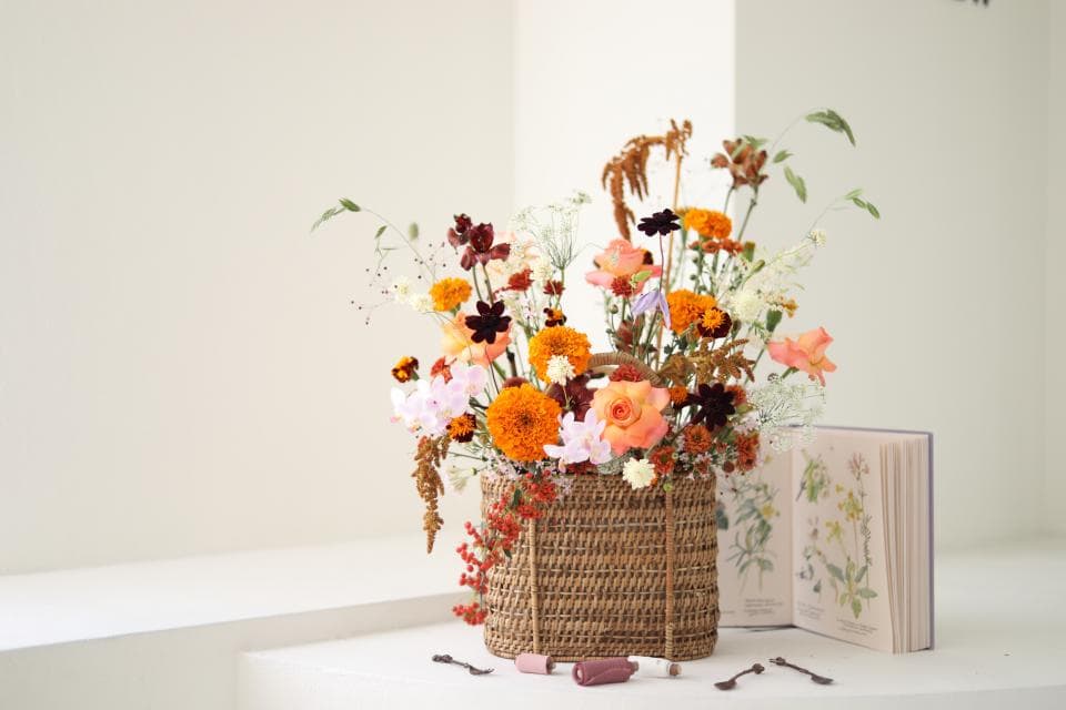 1) 照片顯示一個用藤籃盛裝的秋天花卉,花卉有橙色、黃色、白色和粉色,背景是白色牆壁和一本打開的書籍