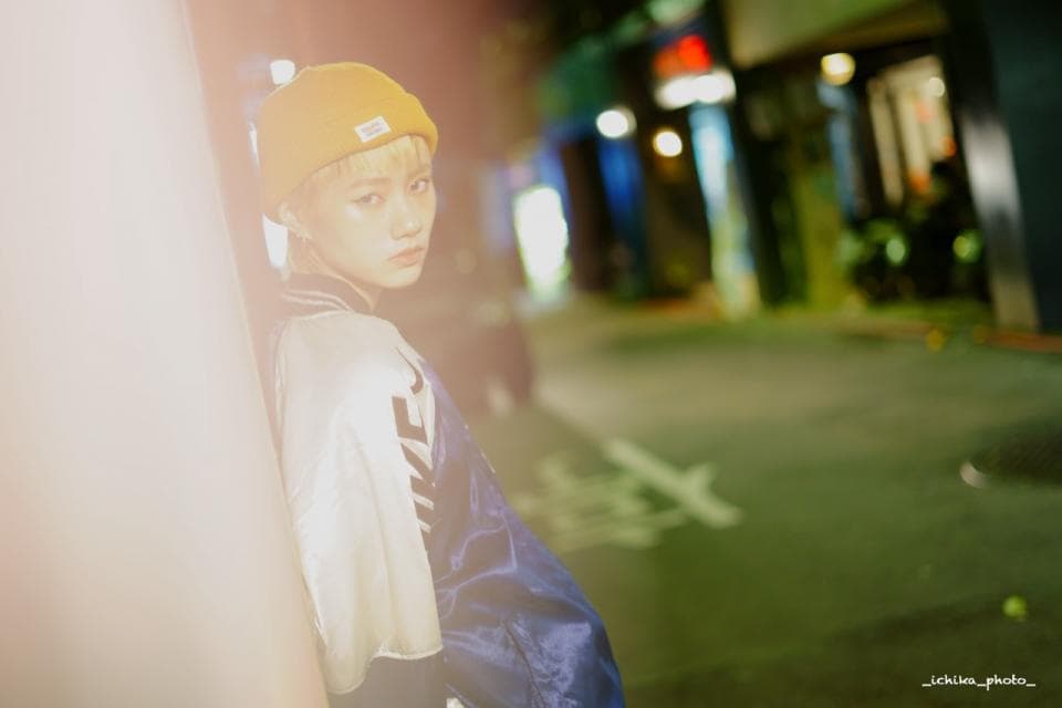 這張照片是一個戴著黃色帽子,穿著藍色外套的年輕人,他側身看著鏡頭,眼神充滿著自信和堅定。他身處在一個夜晚的城市街道,街燈的光芒照亮了他的臉龐,背景是模糊的街道和建築物。