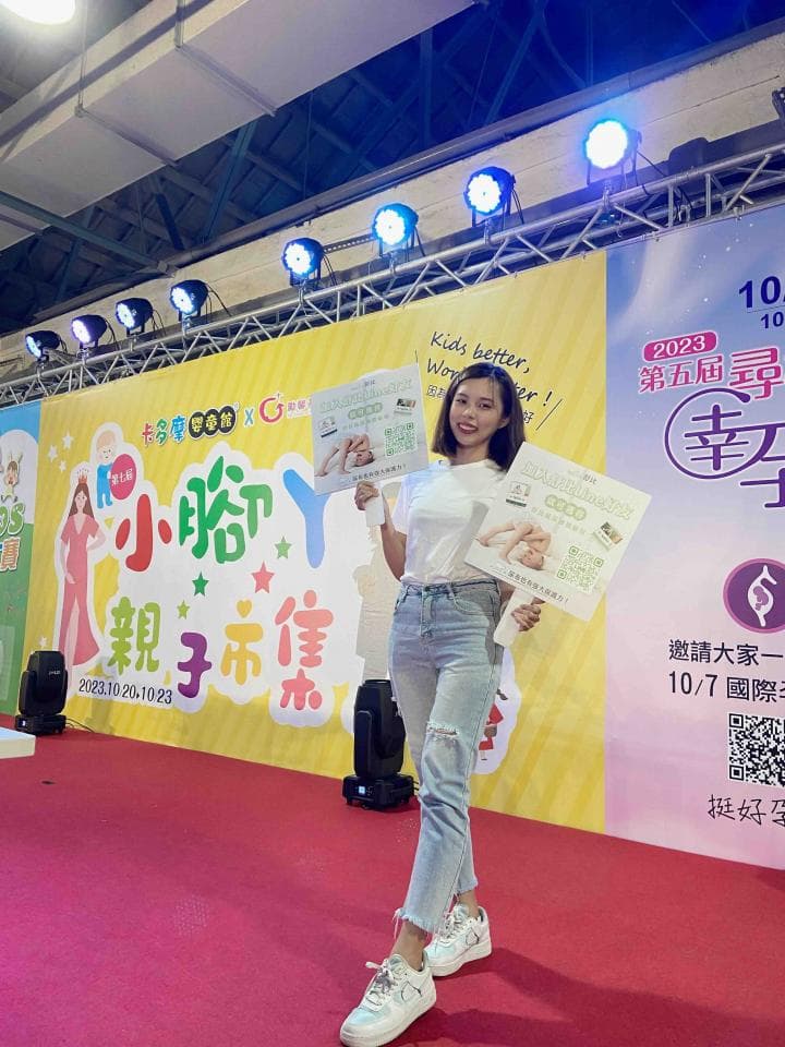 這張照片是一名年輕女性站在舞臺上,她穿著牛仔褲和白色上衣,手裡拿著兩個海報。舞臺背景是黃色的,上面寫著「小小瑜樂子」的字樣。