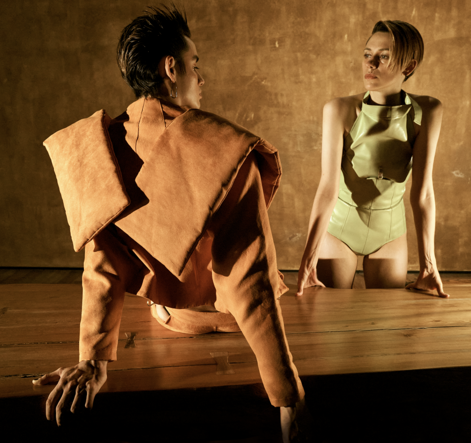 這張照片是一個穿著橘色外套的年輕人,他正坐在一張木桌上,左手放在桌上,右手則放在椅背上。