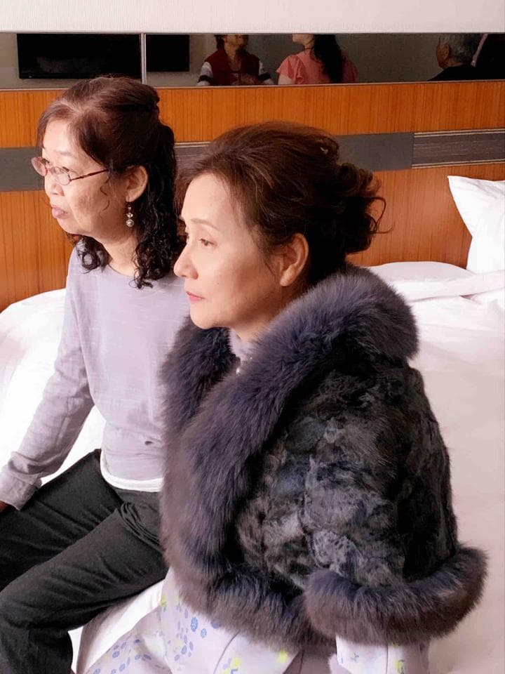 這張「兩個女人坐在床邊」的照片是,一個穿著毛皮外套的女人,旁邊坐著一個穿著毛衣的女人。