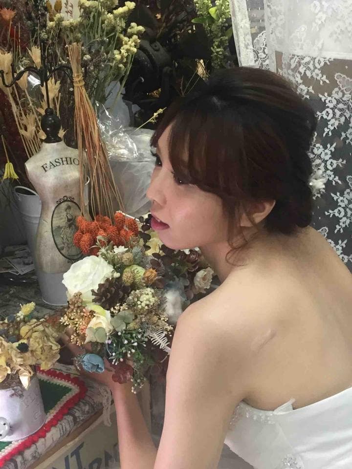 這張「一位女性手拿花束」的照片是,她穿著白色禮服,臉上露出幸福的微笑,背景是花卉和裝飾品。