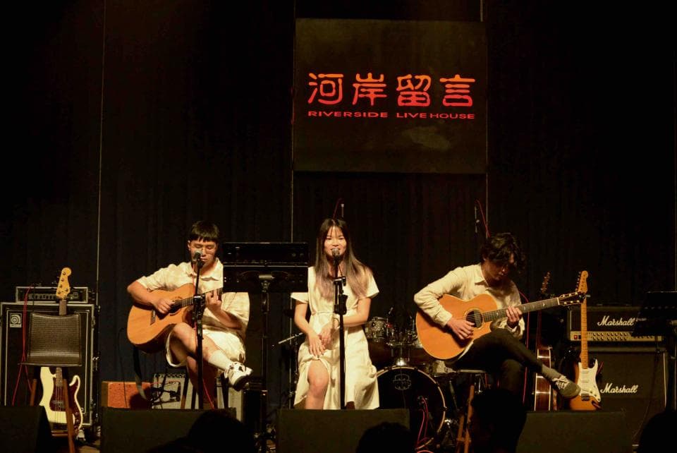 這張照片是三個年輕人在舞臺上演出,中間的女歌手穿著白色連衣裙,正在唱歌,她旁邊的兩個樂手正在演奏吉他。