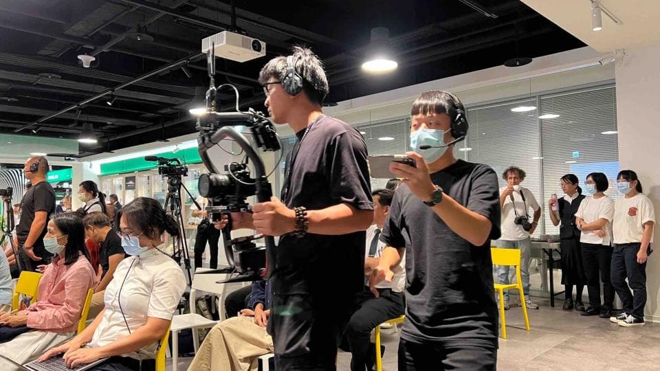 1) 照片中一群人站在房間裡,其中一人手持攝影機,另一人戴著口罩,手持手機