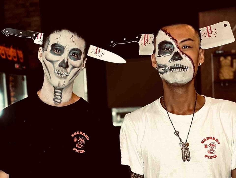 1) 兩位年輕男子,臉上都畫著骷髏頭妝,頭上都戴著刀子,穿著衣服上都有 “Badshah's Pizza” 字樣