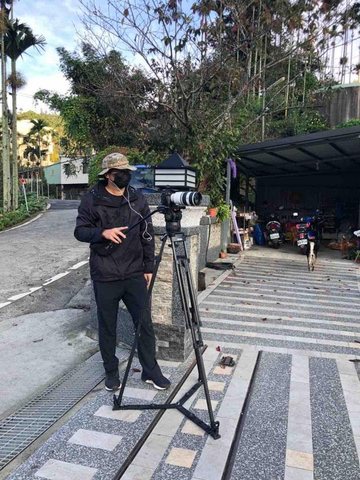 1. 照片中有一名戴著帽子和口罩的男子,他站在一台攝影機前,攝影機架在三腳架上,背景是綠色的樹木和一棟建築物