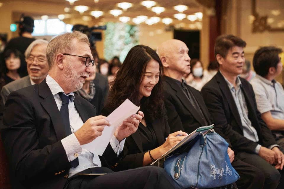 這張「一群人聚會」的照片是,裡面有四位主要的成員:一位穿著西裝的男士正在說話,一位女性正專注地聆聽,而另外兩位則坐在他們身後,其中一位正看著鏡頭,而另一位正低頭沉思。