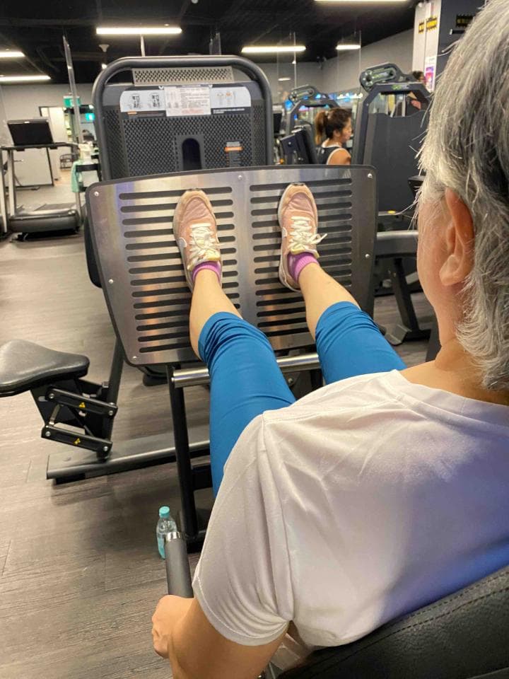 這張照片是一個女人在健身房做腿部運動。她坐在一臺腿部訓練機上,雙腳踏在踏板上,雙手握住把手。她的身體稍微向後傾斜,看起來很專注地做著運動。