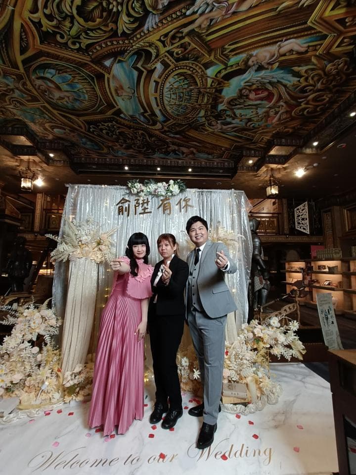 這張「婚禮」的照片是 一對新人和他們的伴娘站在花藝裝飾的婚禮佈置前,背景是華麗的巴洛克式天花板。