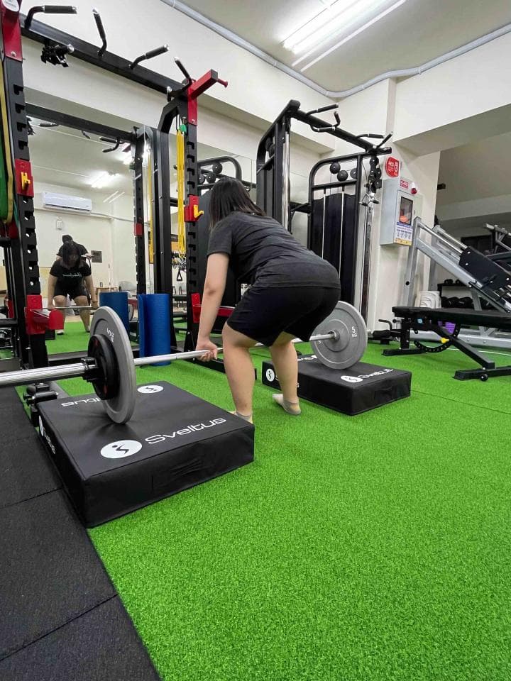 這張照片是一個女子在健身房裡舉重。她站在一個黑色墊子上,雙手握著一根槓鈴,正準備舉起槓鈴。她的表情專注,動作流暢,顯示出她對健身的熱情和努力。