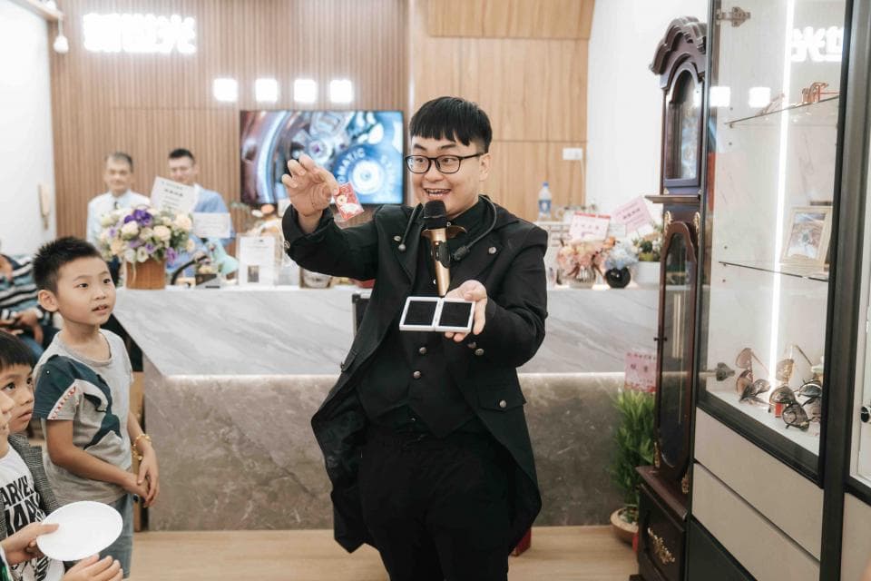 這張照片是一個魔術師正在表演魔術。他穿著黑色的西裝,戴著眼鏡,看起來非常自信。他的手中拿著兩張白色的卡片,正準備表演一個神奇的魔術。