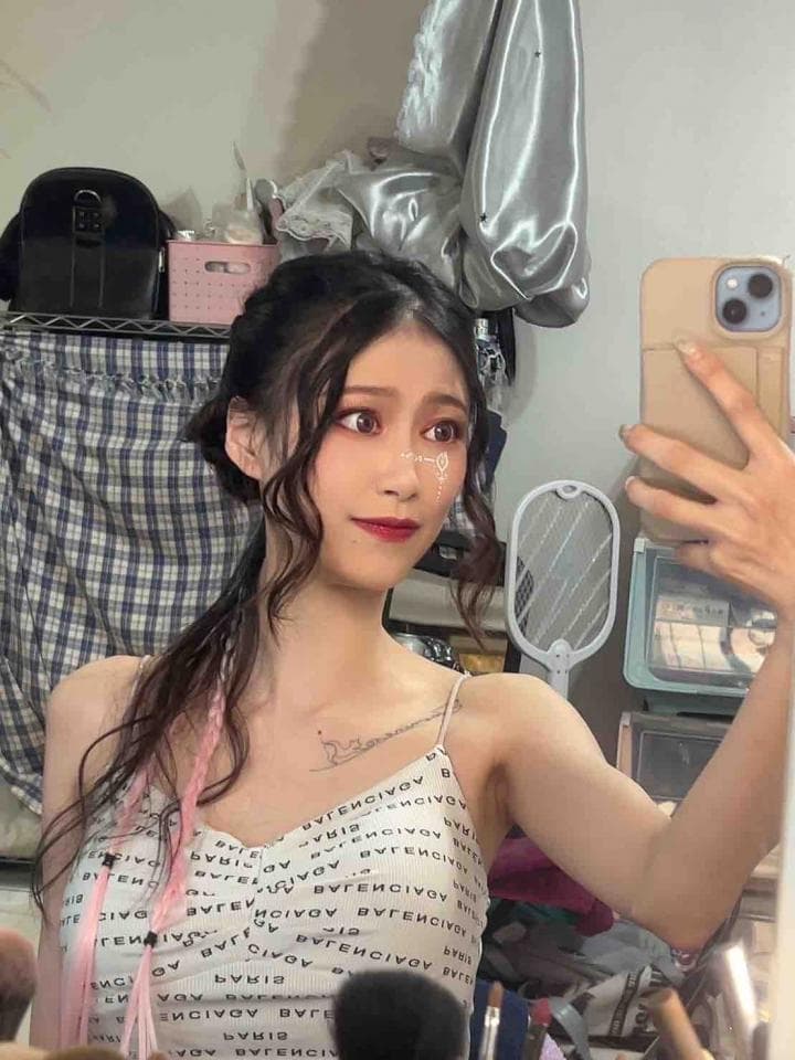 這張照片是一個年輕女孩正在自拍,她穿著一件白色印花背心,頭髮披散在肩上,背景是一些雜亂的物品。