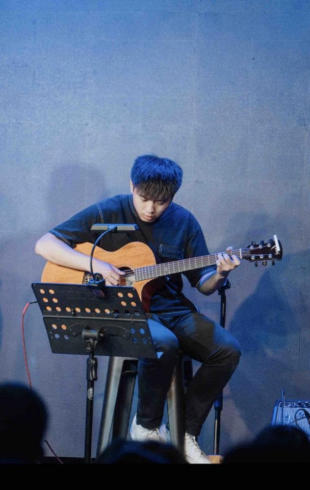 這張照片是一個年輕男子在舞臺上彈吉他,他專注地看著吉他,手指靈活地撥動琴絃,演奏出一段優美的旋律。他的表情認真,似乎沉浸在音樂的世界中。