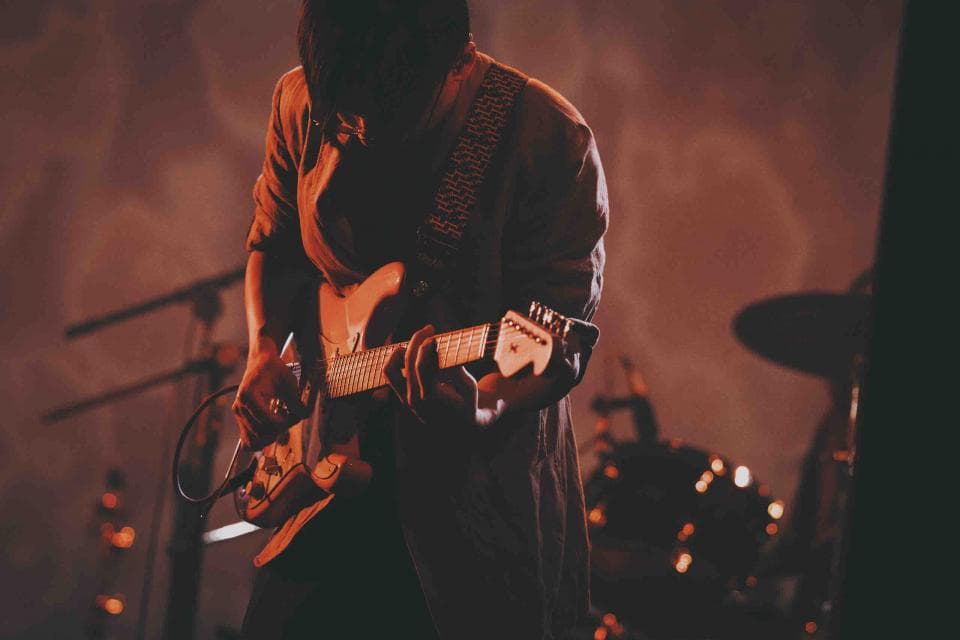 這張照片是一個吉他手在舞台上演奏。他戴著一頂帽子,穿著一件夾克,正在彈奏一把電吉他。他看起來很專注於演奏,音樂似乎充滿了他。