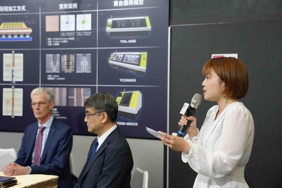 這張照片是一位女士正在臺上用麥克風演講,她身穿白色襯衫,神情專注,手握麥克風,正在演講。