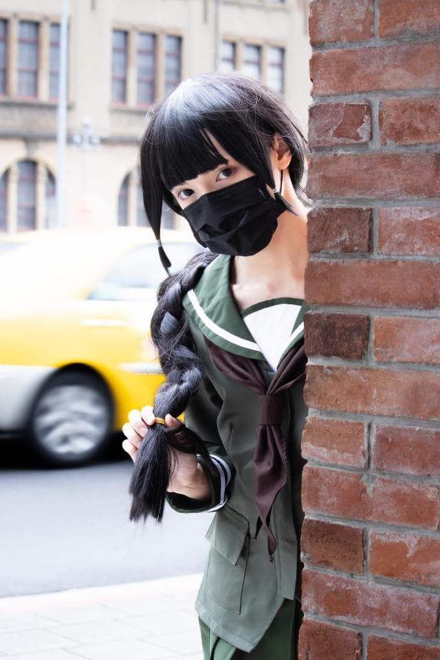 這張「一位戴著口罩的女生,站在紅磚牆邊,她穿著綠色的制服,並用手指梳理著長髮」的照片是。