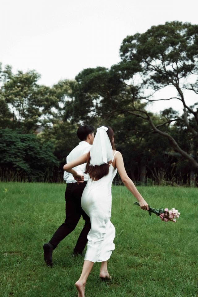 這張「婚紗照」的照片是一對新人,新娘穿著白色婚紗,新郎穿著黑色西裝,他們在草地上奔跑,新娘手持捧花,背景是綠色的樹林。