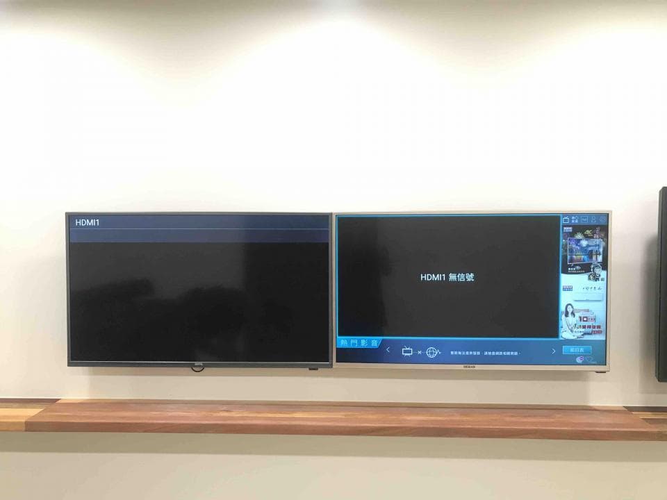 1) 照片顯示兩個電視機,一個是黑色,另一個是銀色的,兩個電視機都掛在牆上,在電視機下有一個木架子