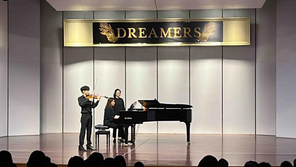 這張照片是一場音樂會,舞台上有一位小提琴手和一位鋼琴家正在演奏。鋼琴家坐在鋼琴前,小提琴手站在鋼琴的左側。舞台上還有一位女性坐在鋼琴旁,可能正在伴奏。舞台背景上有一個金色的牌匾,上面寫着「DREAMERS」,可能是這場音樂會的主題。